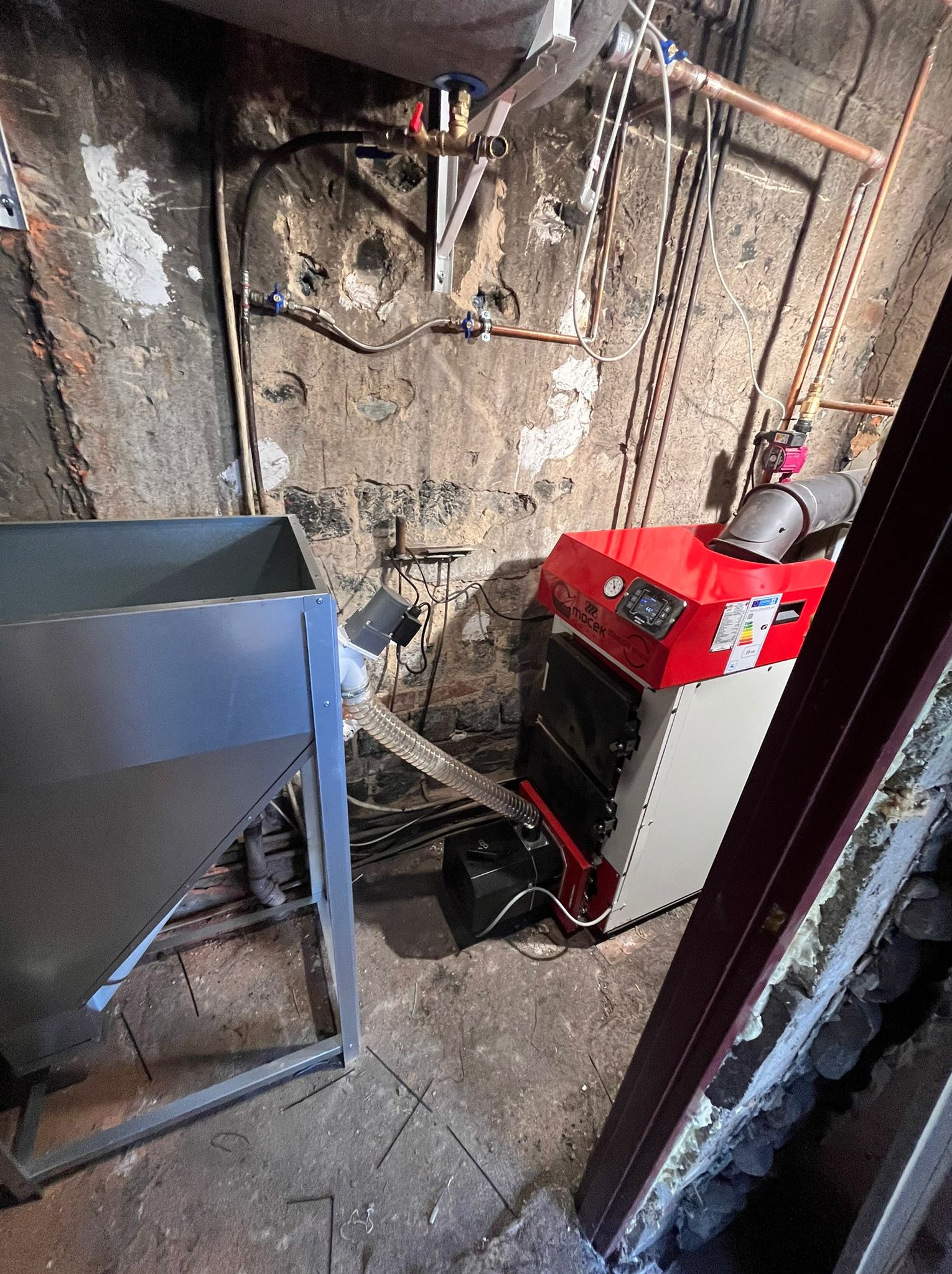 Czerwono-biały piec CO marki Macek w kotłowni w Wysogotowie, z widocznymi rurami instalacji i zbiornikiem na paliwo. Ściany z elementami starej cegły.