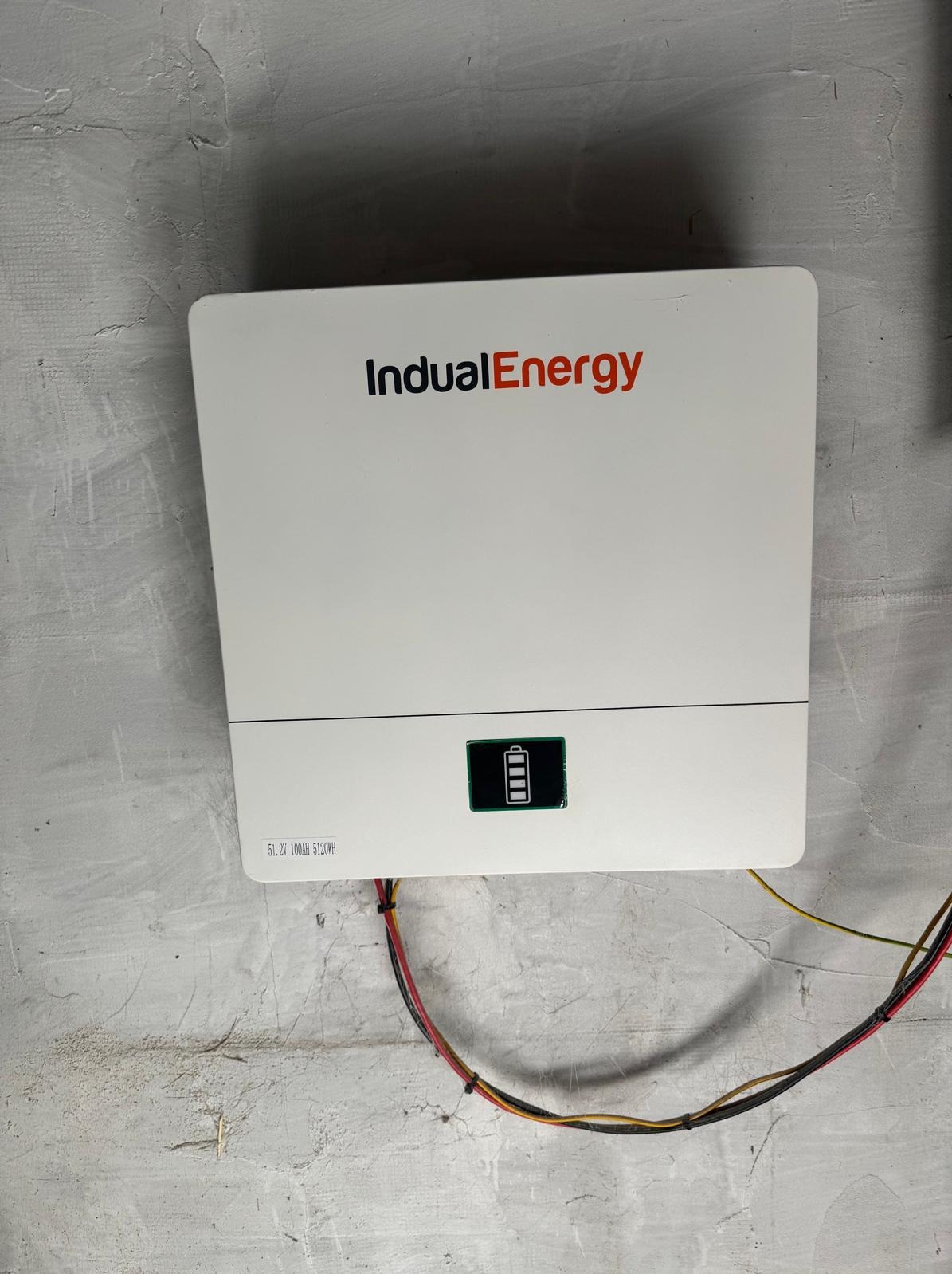 Biały magazyn energii IndualEnergy zamontowany na szarej, surowej ścianie. Widoczne kable i wskaźnik naładowania baterii.