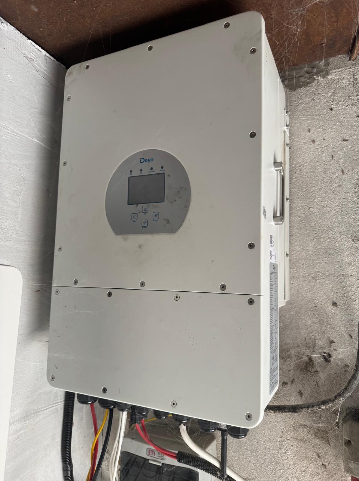 Zamontowany inwerter fotowoltaiczny Deye na szarej ścianie. Widoczne kable i wyświetlacz. Ściana lekko zabrudzona, z pajęczynami w rogu.