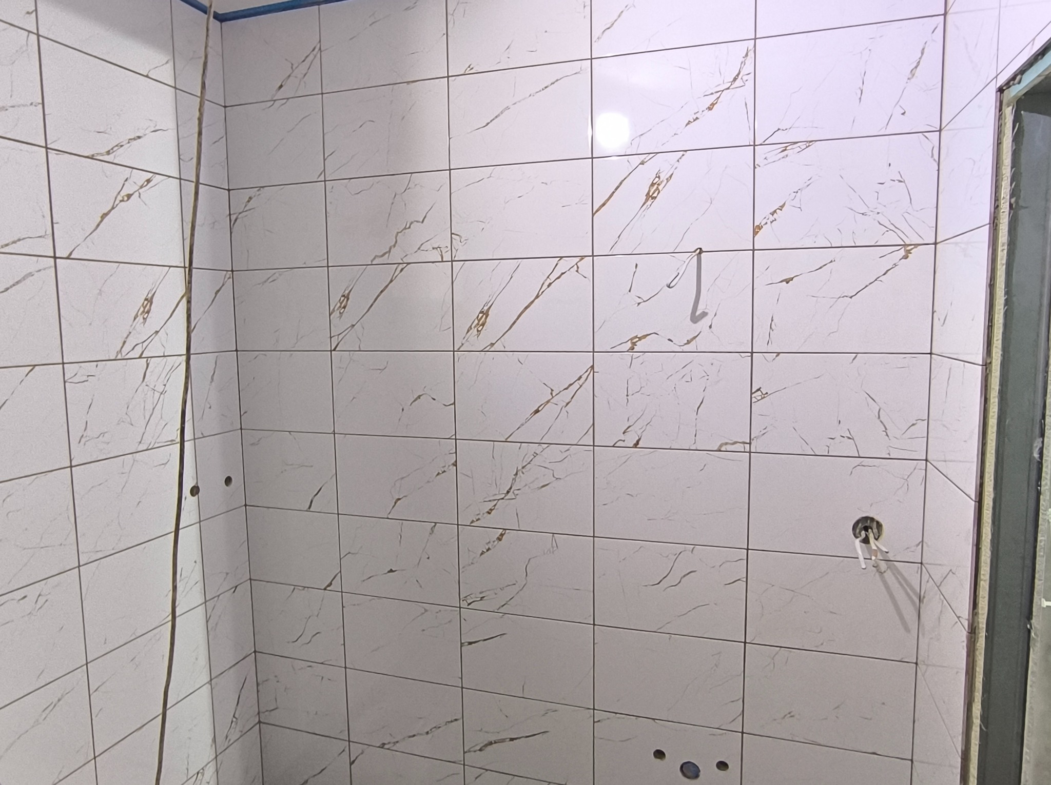 Ściana wyłożona białymi, błyszczącymi płytkami imitującymi marmur z delikatnymi złotymi żyłkami. Widoczne otwory na instalacje i fragment futryny drzwi.