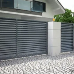 Nowoczesne, szare ogrodzenie lamelowe z betonowymi słupkami na tle domu z balkonem. Kostka brukowa na podjeździe. Model Victory.