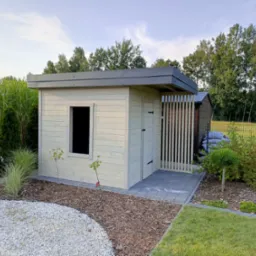 Nowoczesna, jasna drewutnia z płaskim dachem i drewnianą kratownicą wejścia, otoczona zagospodarowanym ogrodem z kamieniami i roślinami ozdobnymi.