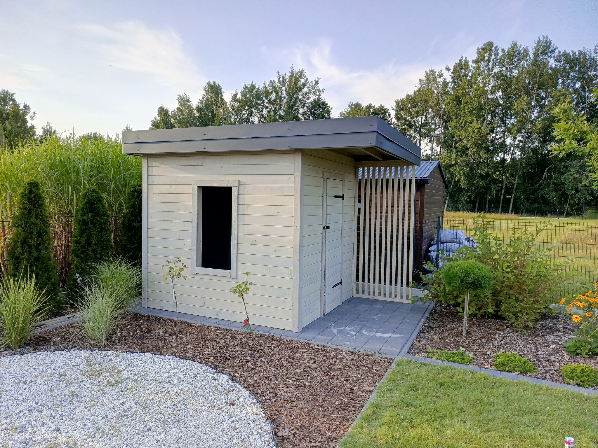 Nowoczesna, jasna drewutnia z płaskim dachem i drewnianą kratownicą wejścia, otoczona zagospodarowanym ogrodem z kamieniami i roślinami ozdobnymi.