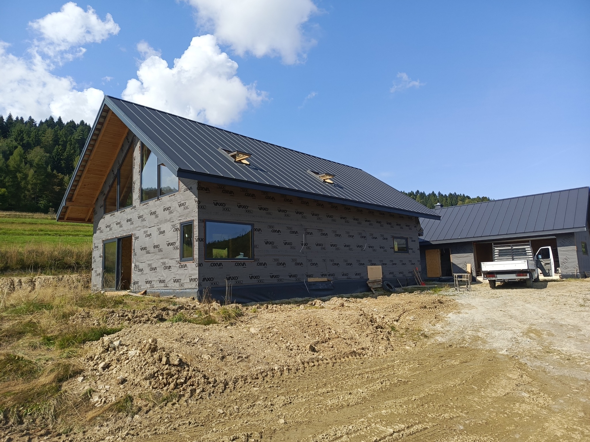 Nowoczesny dom szkieletowy w budowie, z czarnym dachem i folią ochronną na ścianach, na tle błękitnego nieba i zielonego lasu. Widoczny teren budowy i częściowo ukończony budynek gospodarczy.