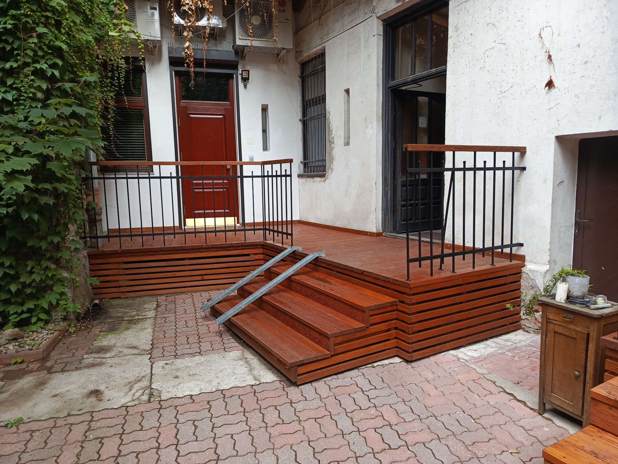 Drewniany taras z balustradą i schodami z poręczami, wykonany przy budynku z elementami zieleni. Czerwona kostka brukowa na podłożu.