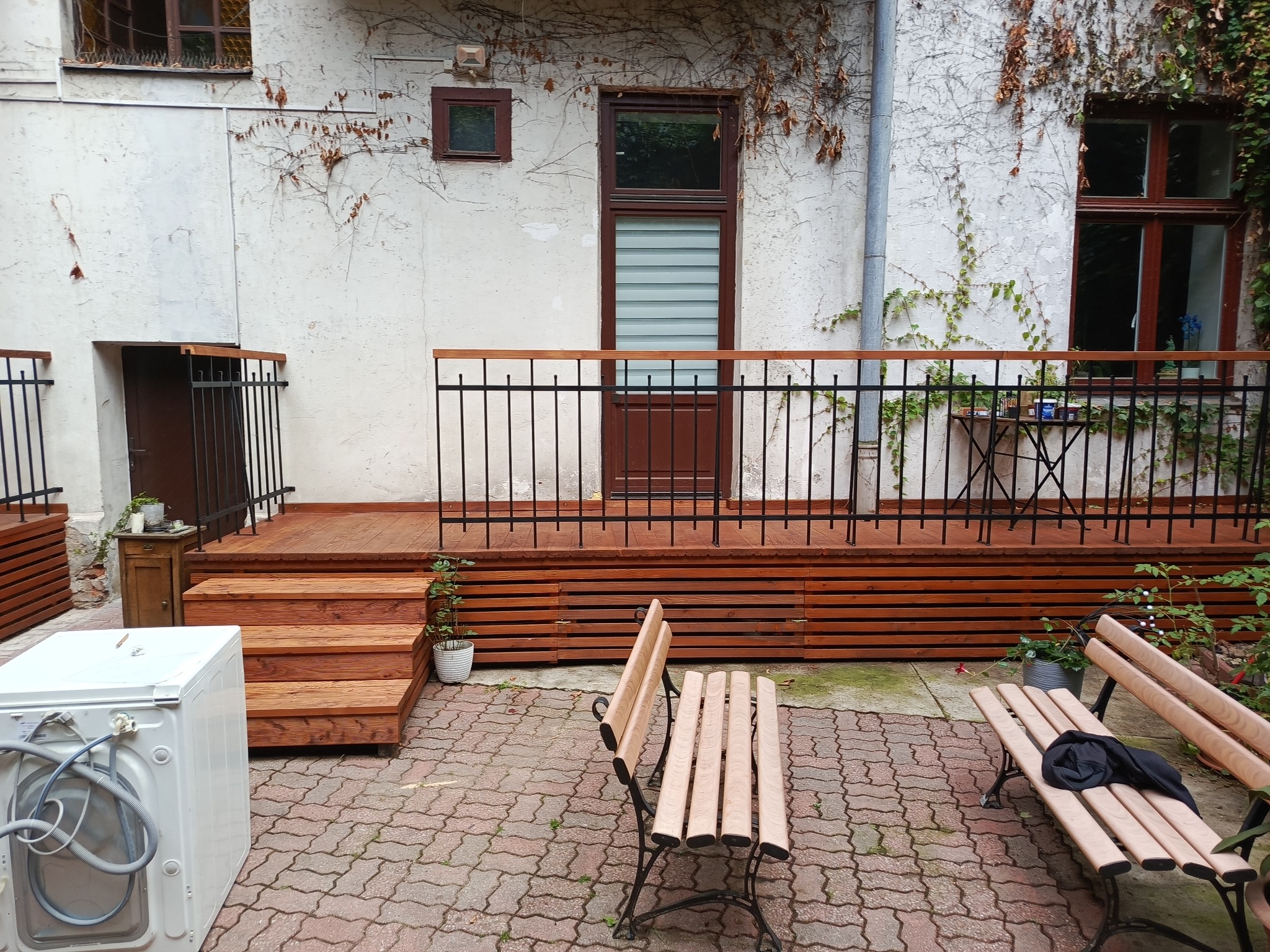 Drewniany taras z czarną balustradą przy budynku z bluszczem. Na pierwszym planie pralka, ławka i kostka brukowa. Nietypowe połączenie przestrzeni użytkowej i rekreacyjnej.