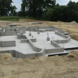 Zalane betonem fundamenty domu jednorodzinnego z wystającymi prętami zbrojeniowymi na działce z ziemią. Widoczne deskowanie i izolacja przeciwwilgociowa.