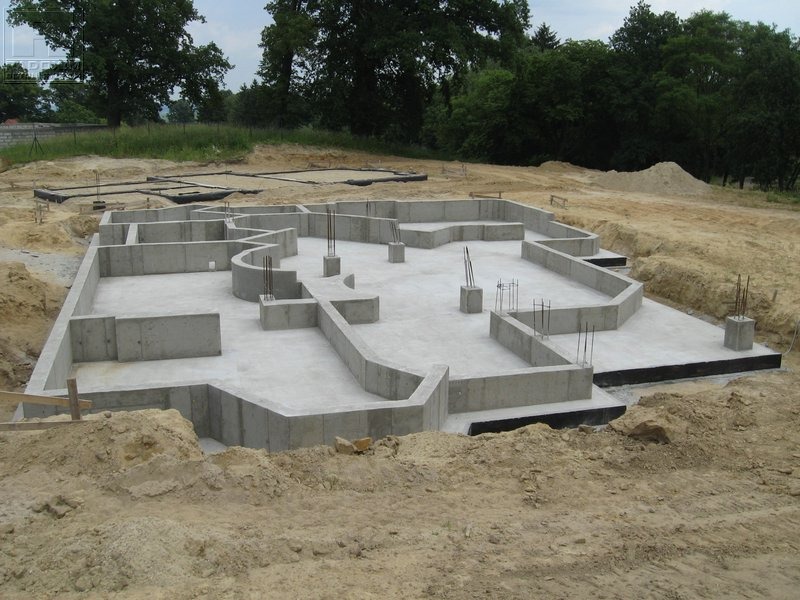 Zalane betonem fundamenty domu jednorodzinnego z wystającymi prętami zbrojeniowymi na działce z ziemią. Widoczne deskowanie i izolacja przeciwwilgociowa.