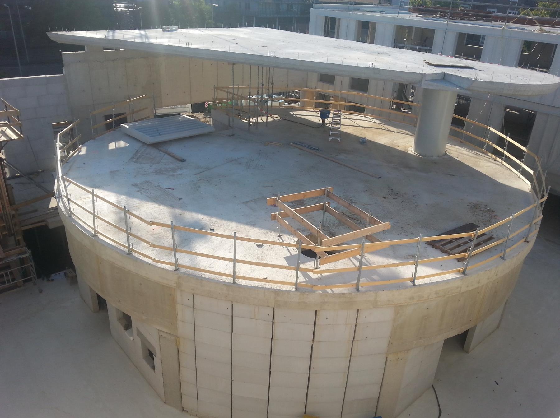 Wysoka perspektywa na okrągły budynek w trakcie budowy z żelbetu. Widoczne szalunki, zbrojenia i częściowo wylane elementy. Jasne oświetlenie.