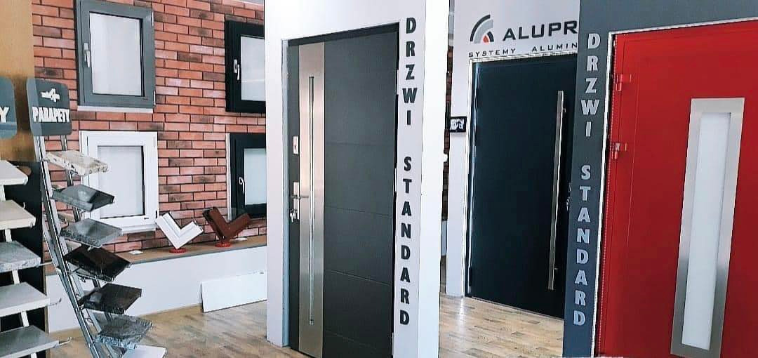 Ekspozycja drzwi i okien w salonie: szare drzwi z metalowym panelem, czarne okna na ceglanej ścianie, czerwone drzwi z białym panelem, okna aluminiowe.