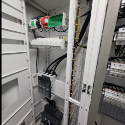 Adam Topolewski - Wnętrze szafy elektrycznej z widocznymi zabezpieczeniami, listwami zaciskowymi i przewodami. Górna część szafy oświetlona zielonym światłem.