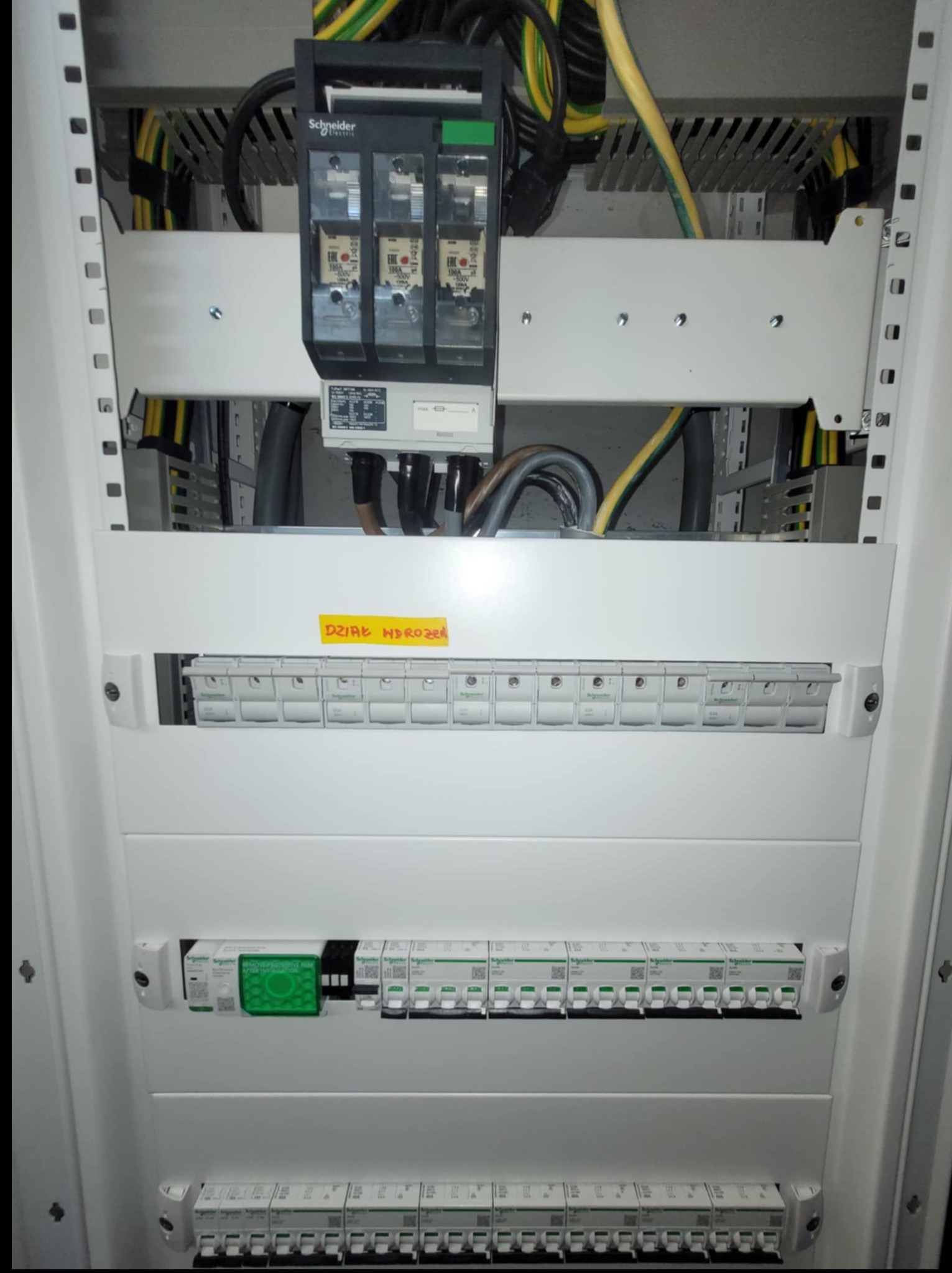 Wnętrze szafy elektrycznej z zainstalowanymi bezpiecznikami, przewodami i aparaturą Schneider Electric. Widoczna tabliczka z napisem 'DZIAŁ HYDROZĘ'.