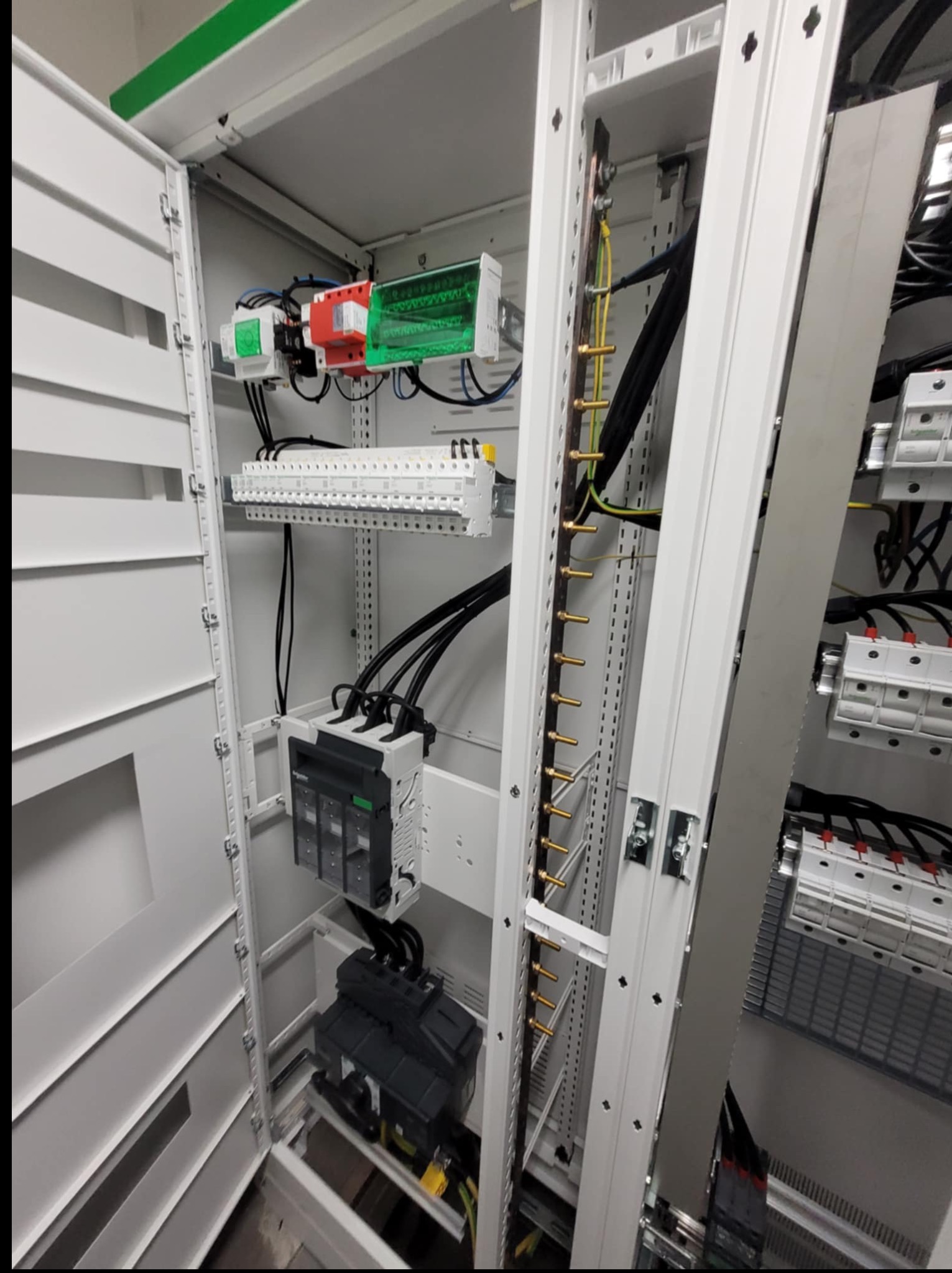 Wnętrze szafy elektrycznej z widocznymi zabezpieczeniami, listwami zaciskowymi i przewodami. Górna część szafy oświetlona zielonym światłem.