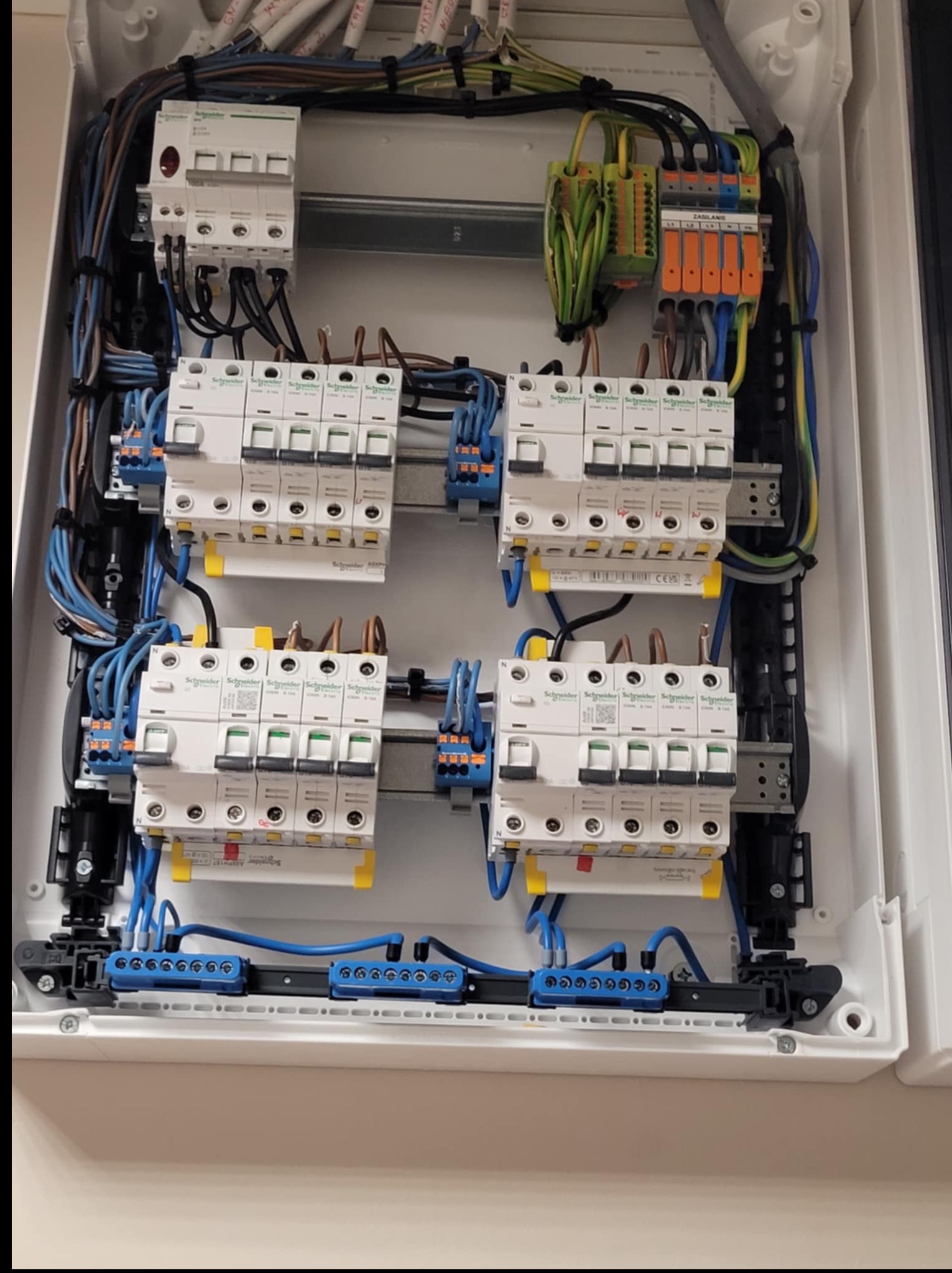 Wewnętrzny widok rozdzielnicy elektrycznej z zainstalowanymi bezpiecznikami Schneider, przewodami o różnych kolorach i złączkami. Widoczne elementy montażowe i okablowanie.