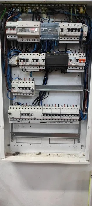Rozdzielnica elektryczna z licznikiem i zabezpieczeniami, widoczne okablowanie. Całość w białej obudowie, zamontowana na ścianie.