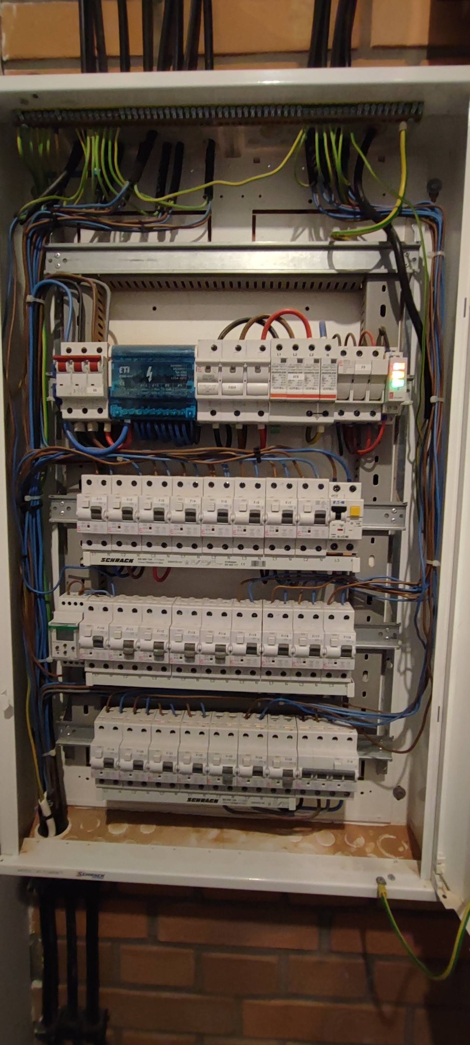 Rozdzielnica elektryczna z zainstalowanymi bezpiecznikami, przewodami i licznikiem, zamontowana na ceglanej ścianie. Widoczne elementy marki Schrack.