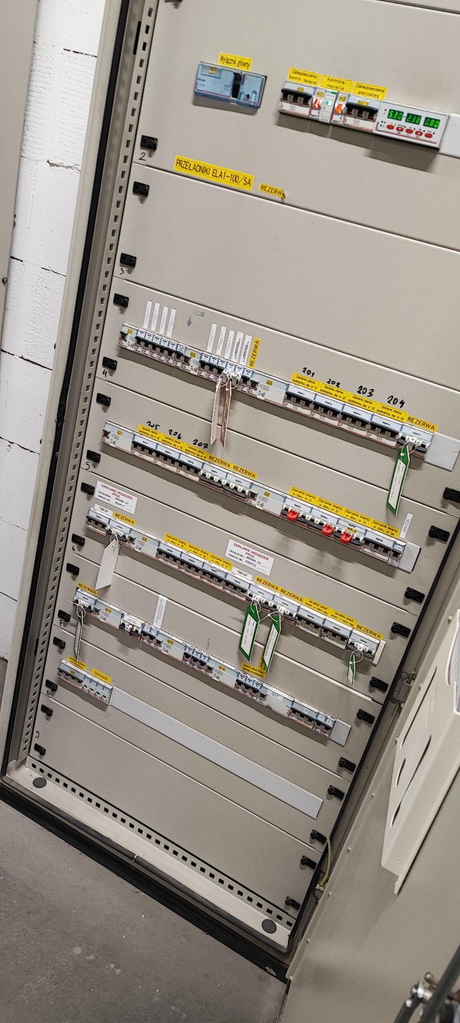 Szara szafa sterownicza z bezpiecznikami, opisana etykietami i numerami. Widoczne zabezpieczenia, wyłączniki i oznaczenia rezerwowe. Element instalacji wewnątrz pomieszczenia.