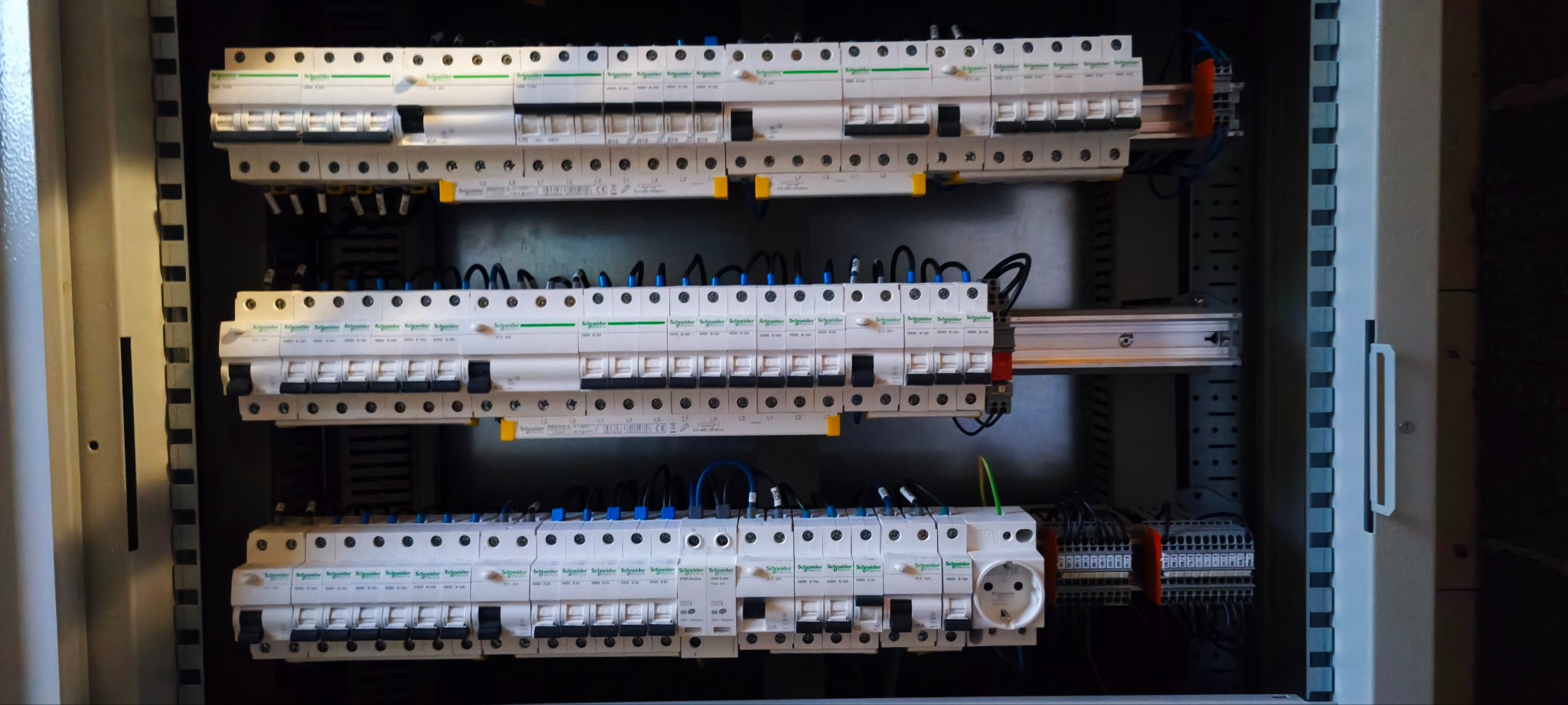 Rozdzielnica elektryczna z zainstalowanymi bezpiecznikami Schneider, widoczne przewody i gniazdo. Elementy zamontowane w metalowej obudowie.