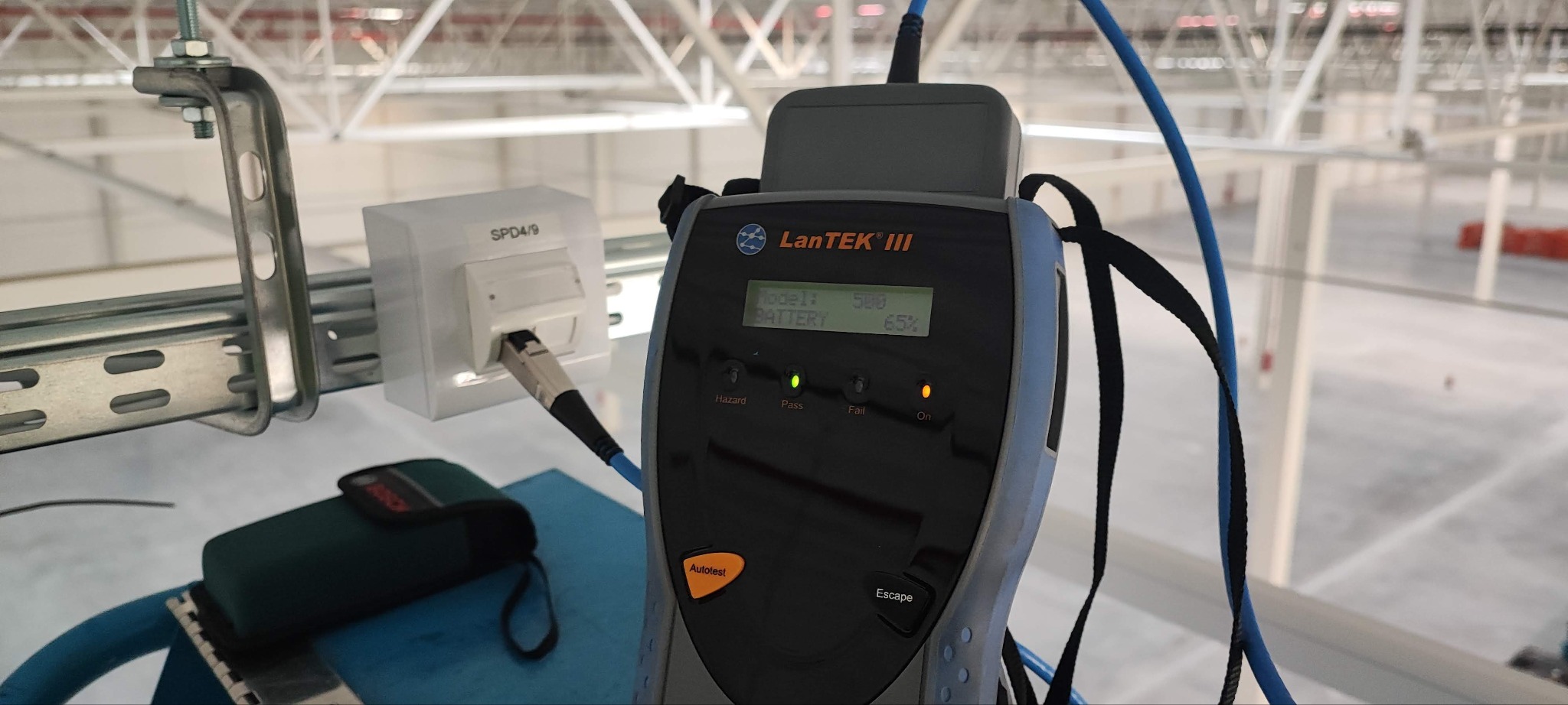 Tester LanTEK III mierzy parametry kabla sieciowego RJ45 w gnieździe. Widoczna zielona dioda 'Pass', tło to hala przemysłowa z konstrukcją wsporczą.