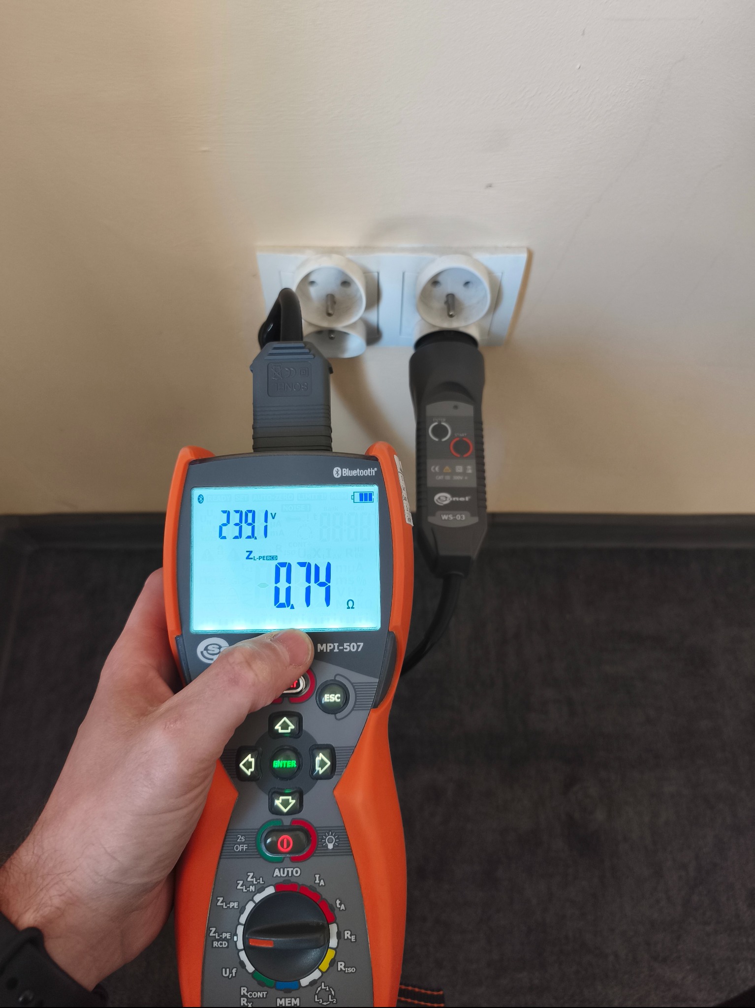 Miernik MPI-507 w trakcie pomiaru napięcia w gniazdku elektrycznym. Na wyświetlaczu widoczne wartości 239.1V i 0.74 Ω. Urządzenie podłączone do gniazdka za pomocą sond.