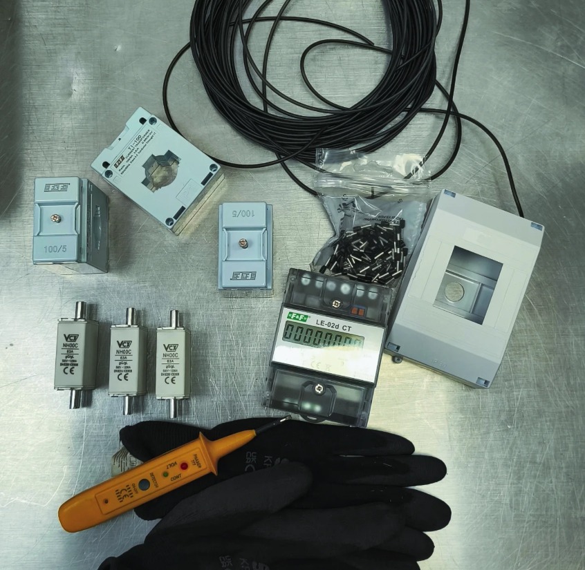 Zestaw komponentów elektrycznych: przekładniki prądowe, bezpieczniki NH, licznik energii, przewód, złączki i próbnik napięcia na metalowym stole, gotowe do instalacji.