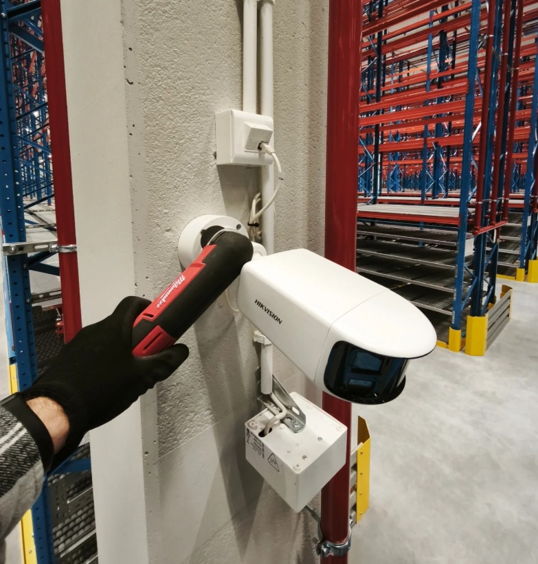 Montaż kamery przemysłowej Hikvision na betonowym słupie w magazynie. Widoczna ręka w rękawicy trzymająca narzędzie Milwaukee.