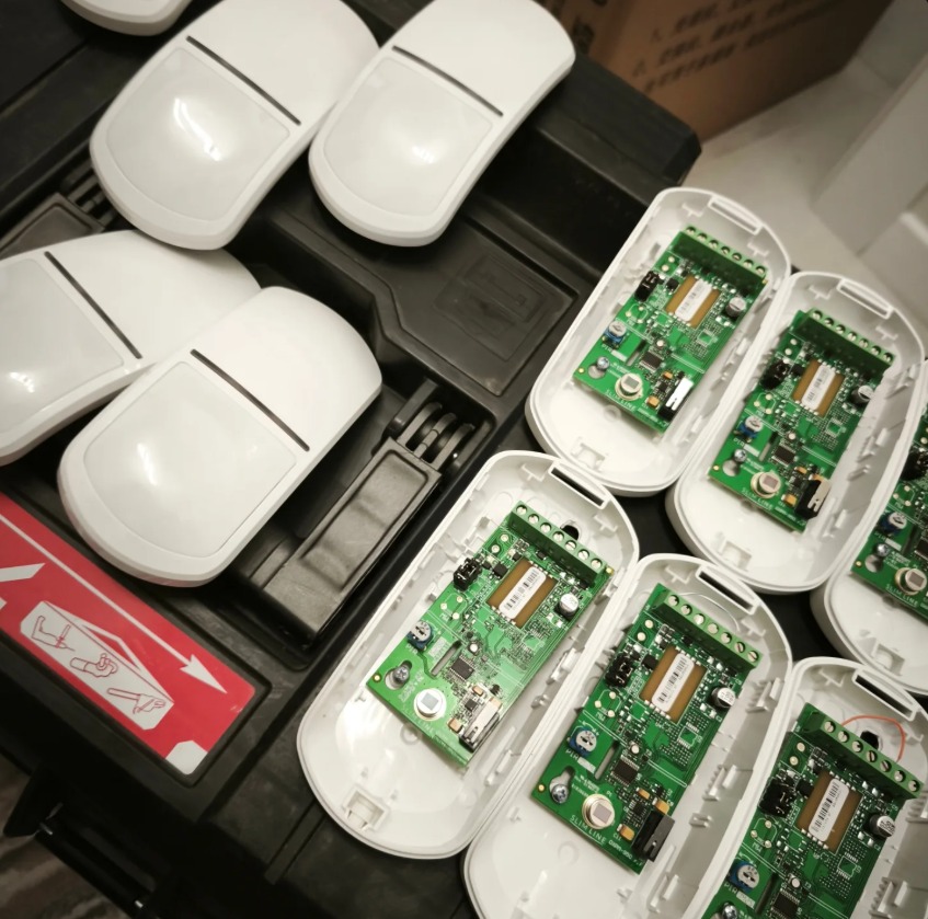 Rozłożone czujniki ruchu z widocznymi płytkami drukowanymi i obudowami na czarnym tle. Widoczny element ostrzegawczy z piktogramem.