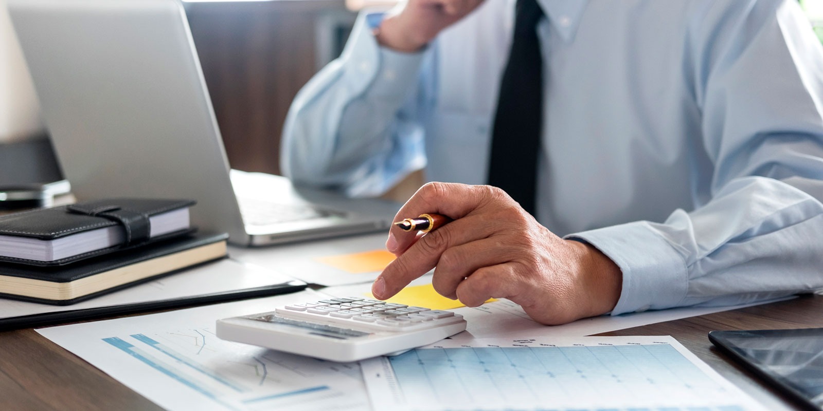 Księgowy przy biurku: kalkulator, laptop, dokumenty. Dłoń z piórem nad klawiaturą kalkulatora. Analiza danych finansowych w biurze rachunkowym.