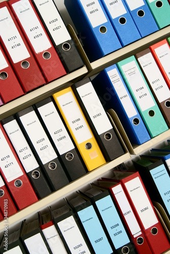 Półki z segregatorami w różnych kolorach, etykiety z numerami, ułożone pod kątem, sugerujące uporządkowaną archiwizację dokumentów w biurze rachunkowym.