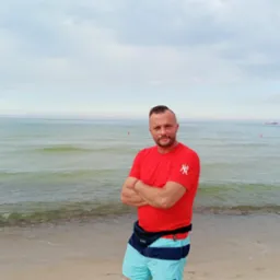 Mężczyzna w czerwonej koszulce z założonymi rękami stoi na piaszczystej plaży z widokiem na morze i pochmurne niebo w tle. Ujęcie centralne, lekko od dołu.