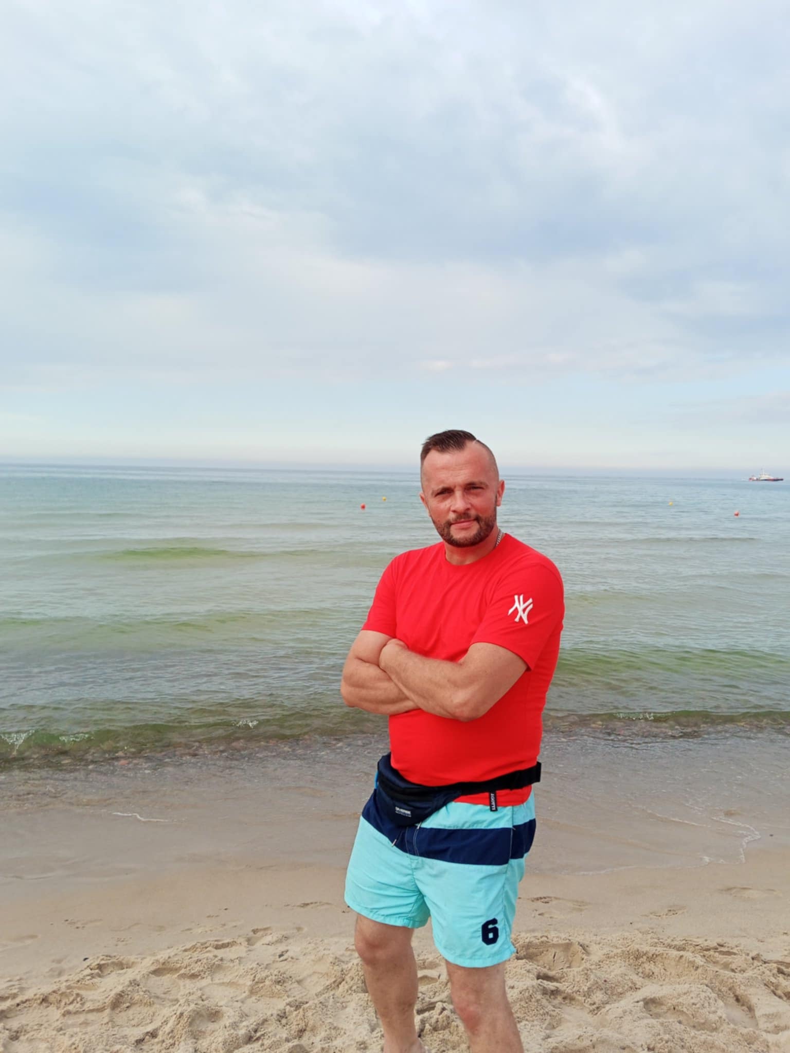 Mężczyzna w czerwonej koszulce z założonymi rękami stoi na piaszczystej plaży z widokiem na morze i pochmurne niebo w tle. Ujęcie centralne, lekko od dołu.
