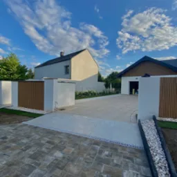 Nowoczesny podjazd z kostki brukowej w odcieniach szarości i beżu, oddzielony od jasnych płyt drenażem liniowym. W tle dom z garażem i fragment ogrodzenia.