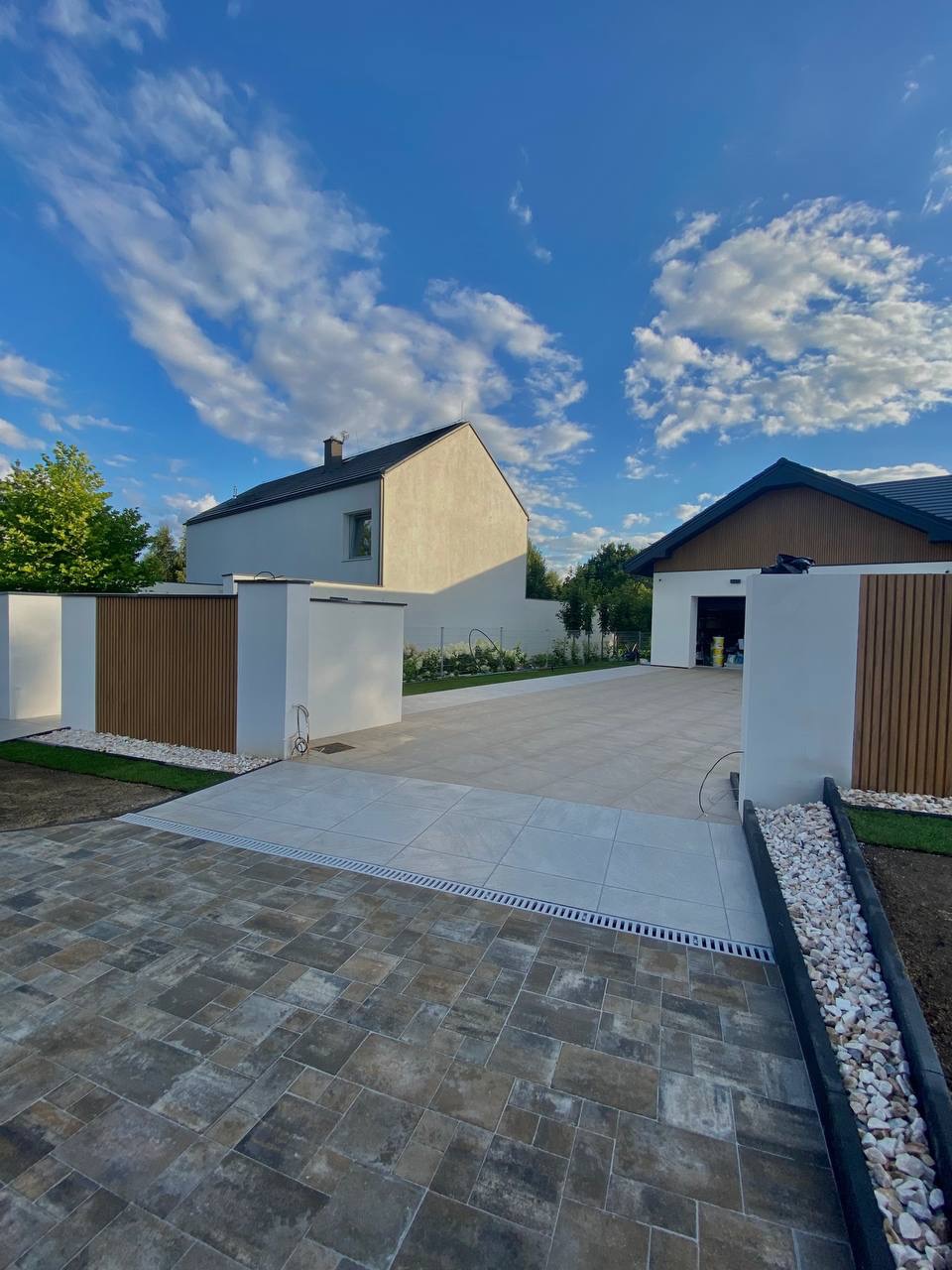 Nowoczesny podjazd z kostki brukowej w odcieniach szarości i beżu, oddzielony od jasnych płyt drenażem liniowym. W tle dom z garażem i fragment ogrodzenia.
