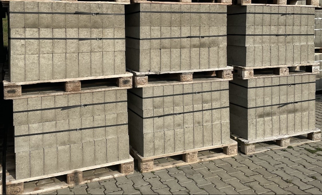 Szare bloczki betonowe ułożone na paletach, zabezpieczone czarną taśmą. Widoczna kostka brukowa w tle. Bloczki o regularnych kształtach, ustawione jeden na drugim.