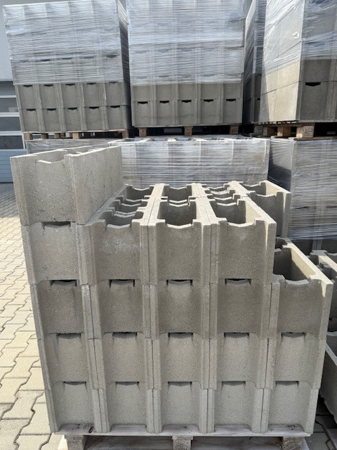 Pustaki szalunkowe ułożone w stos na palecie, widoczne charakterystyczne otwory. W tle kolejne palety z pustakami zabezpieczone folią.