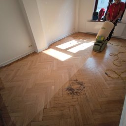 Wykończenia Wnętrz Małgorzata Szuchalska - Parkiet w jodełkę po częściowym cyklinowaniu. Widoczne różnice w kolorze drewna, maszyna do cyklinowania w tle, osoba obsługująca maszynę. Oświetlenie dzienne.