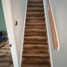 Wykończenia Wnętrz Małgorzata Szuchalska - Schody wykończone panelami drewnopodobnymi z białą listwą, widok z dołu. Ściany szare, biała balustrada. Fragment podłogi i narzędzi widoczny w tle.