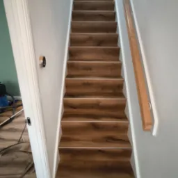 Schody wykończone panelami drewnopodobnymi z białą listwą, widok z dołu. Ściany szare, biała balustrada. Fragment podłogi i narzędzi widoczny w tle.