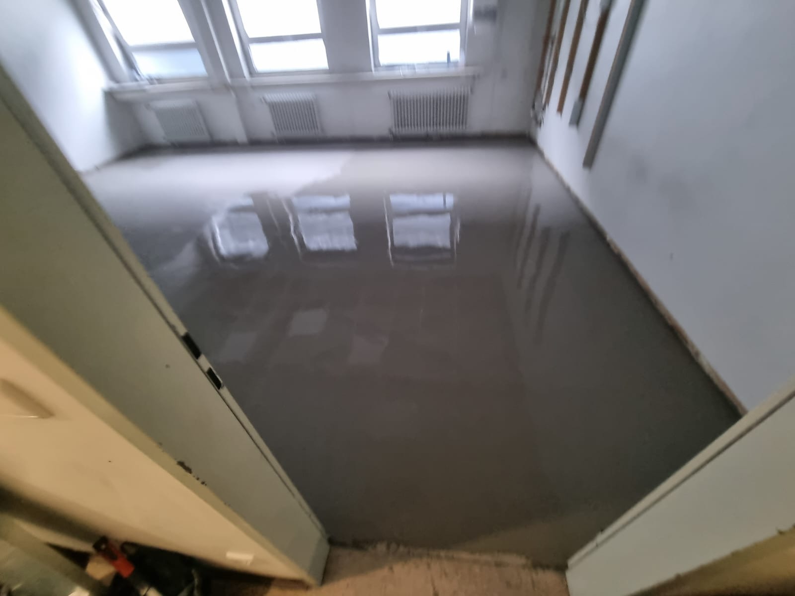 Wylana, świeża wylewka samopoziomująca w jasnym pokoju z oknami dachowymi. Widok zza futryny drzwi. Na ścianach widoczne listwy przypodłogowe.