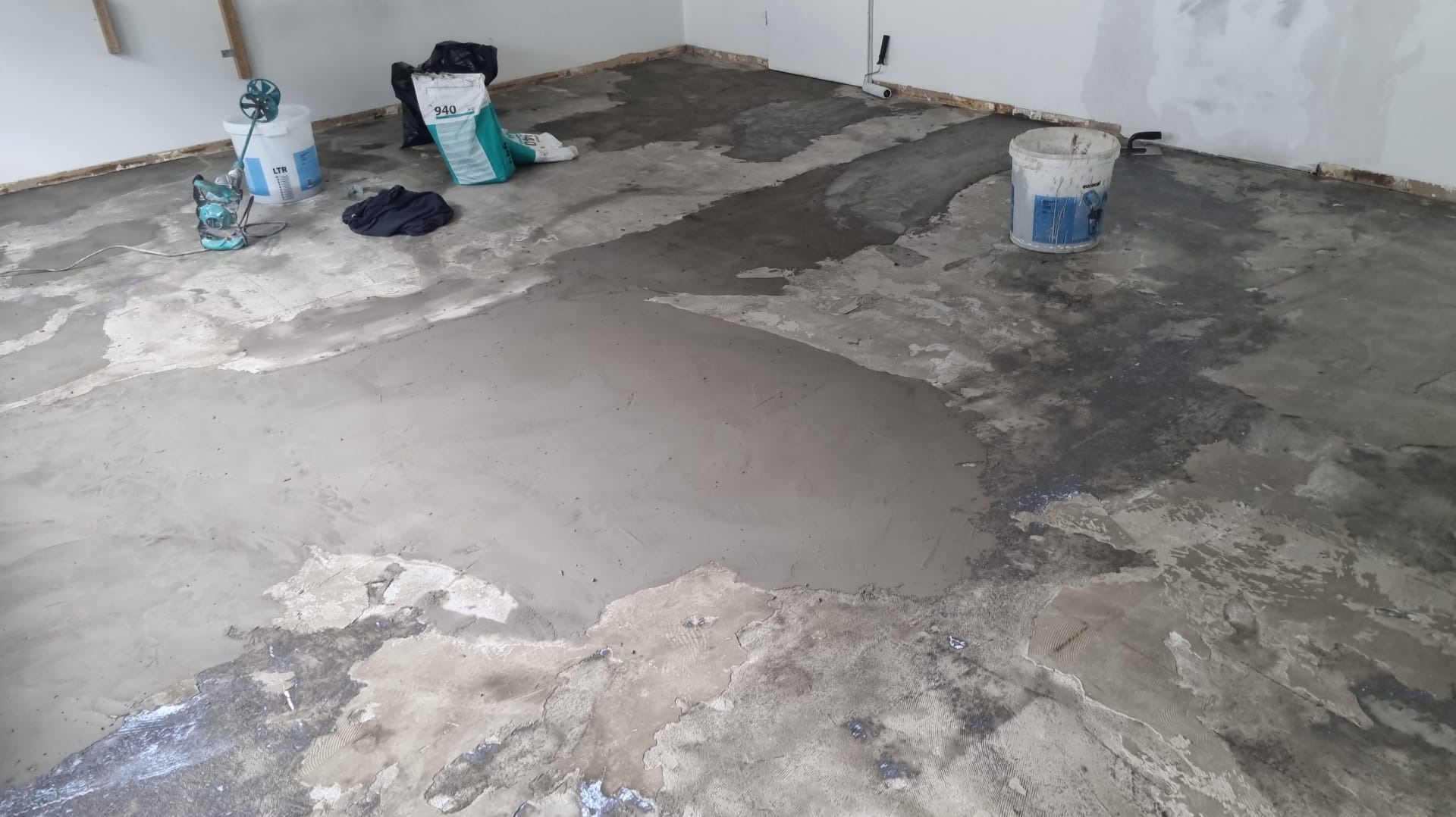 Wyrównywanie podłoża w pomieszczeniu szpachlą. Widoczne narzędzia, wiadro, worek z zaprawą. Częściowo wykończona podłoga z widocznymi śladami wcześniejszych warstw.