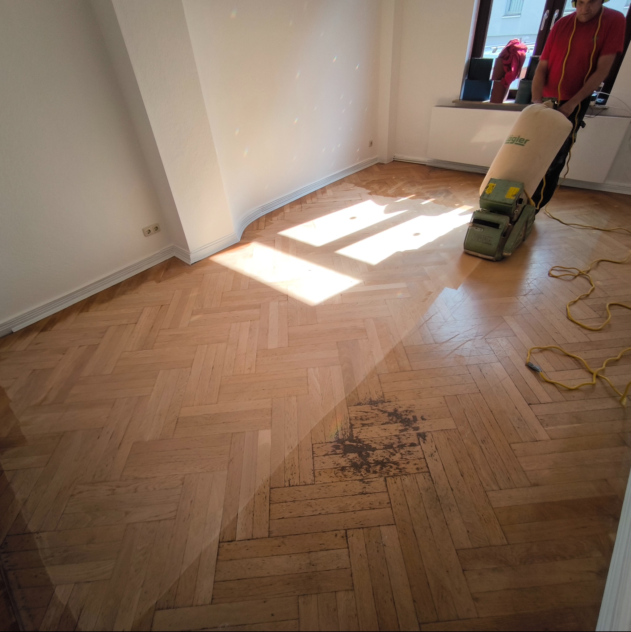 Parkiet w jodełkę po częściowym cyklinowaniu. Widoczne różnice w kolorze drewna, maszyna do cyklinowania w tle, osoba obsługująca maszynę. Oświetlenie dzienne.