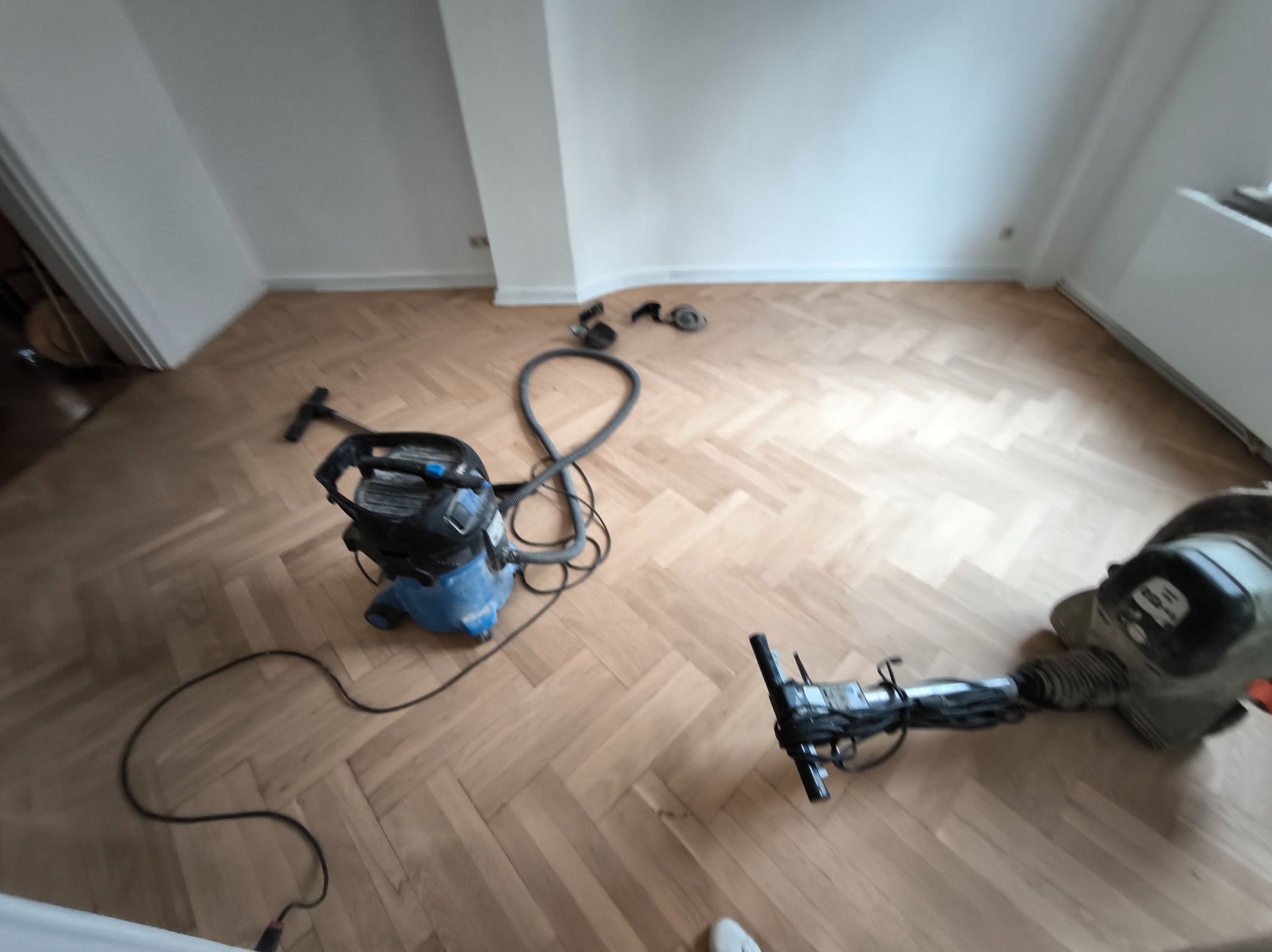 Parkiet w jodełkę po cyklinowaniu. Widoczne maszyny: cykliniarka i odkurzacz przemysłowy. Świeżo wycyklinowana, jasna podłoga w pustym pokoju.