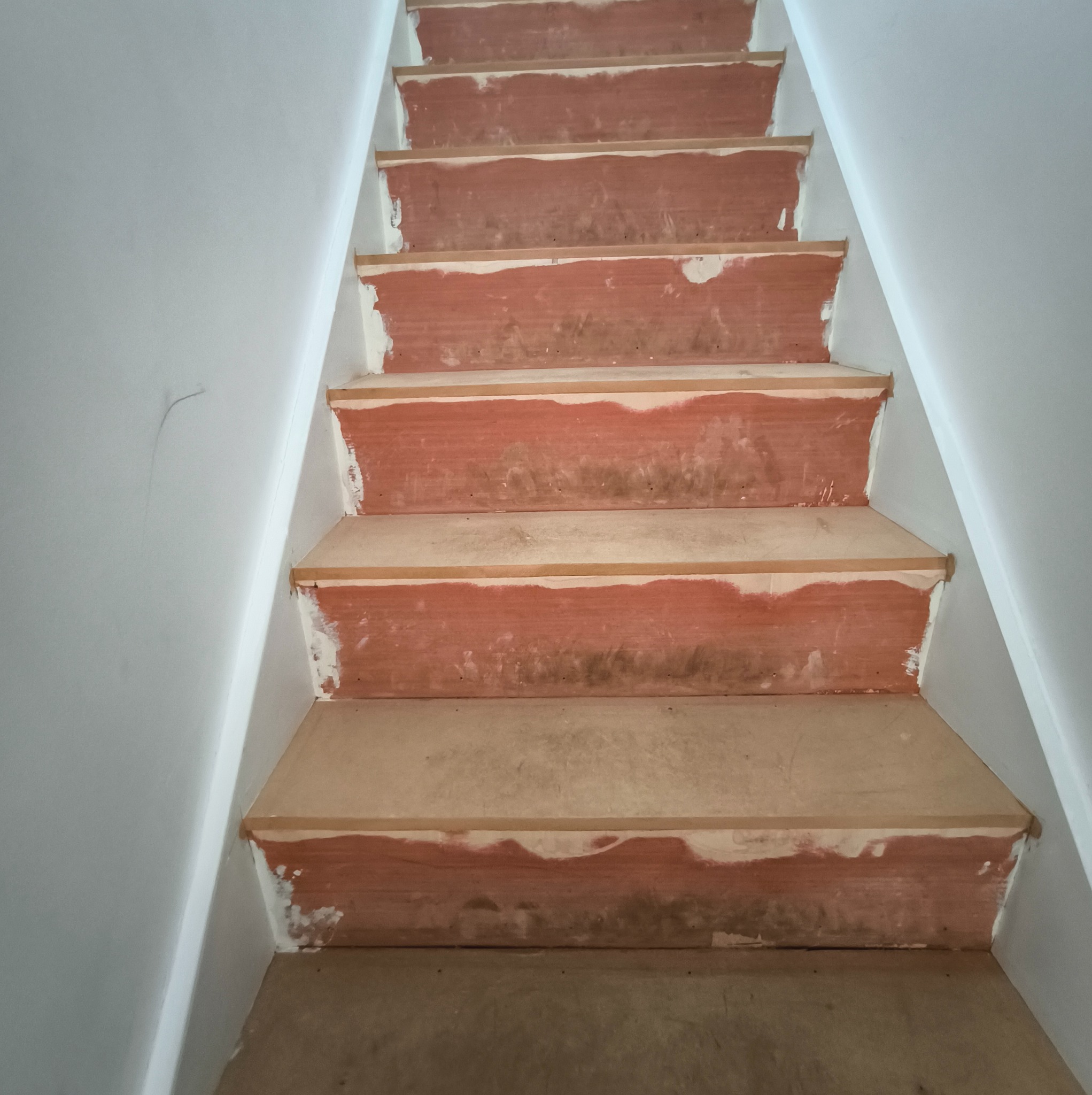 Drewniane schody w trakcie renowacji, z widocznymi śladami po szpachlowaniu i przygotowaniu do malowania, na tle białej ściany. Ujęcie z dołu schodów.