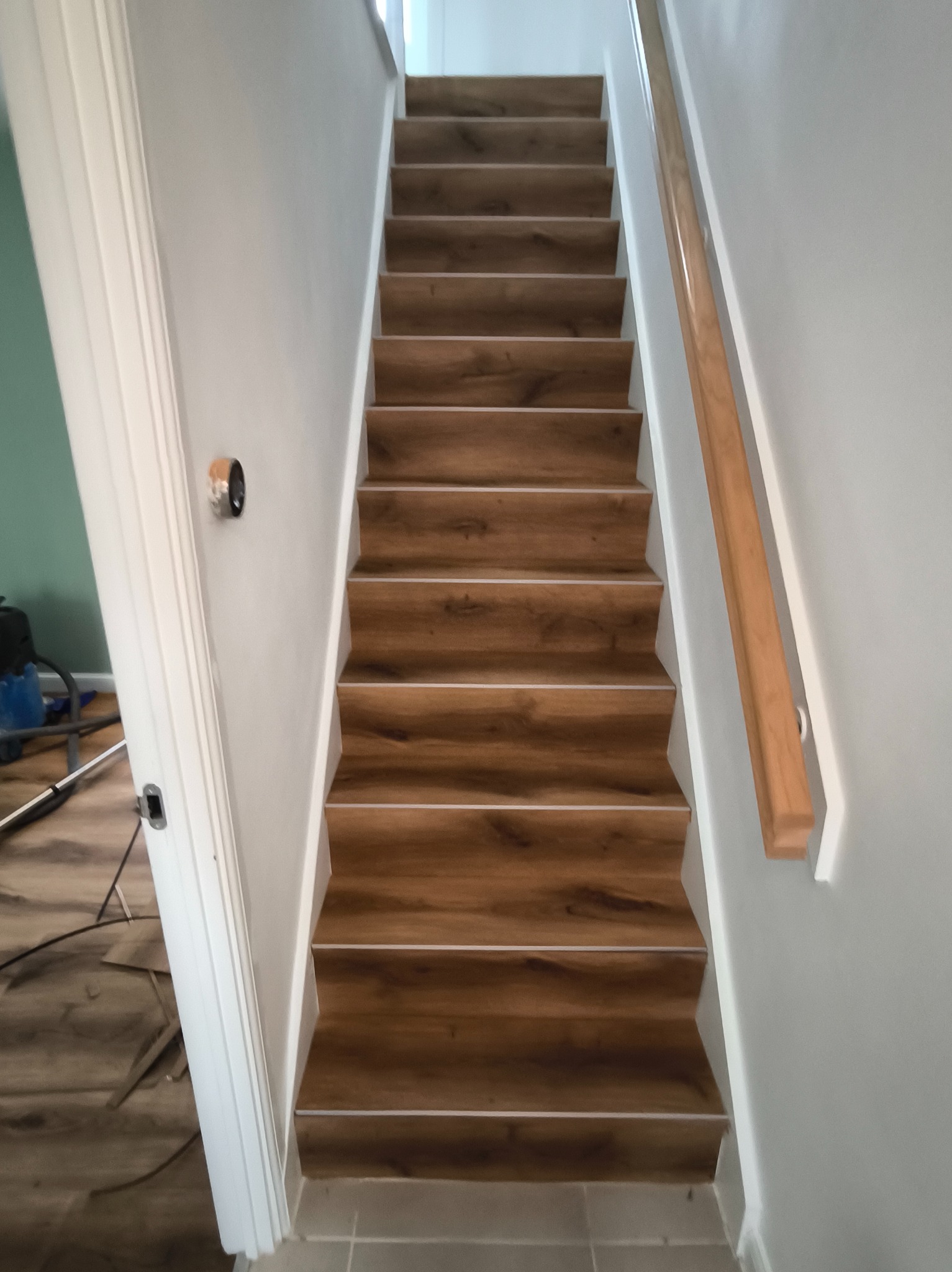 Schody wykończone panelami drewnopodobnymi z białą listwą, widok z dołu. Ściany szare, biała balustrada. Fragment podłogi i narzędzi widoczny w tle.