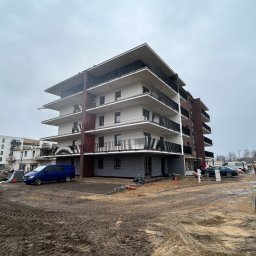 IZBUD - Wielopiętrowy budynek mieszkalny z balkonami w trakcie prac wykończeniowych, widoczny sprzęt budowlany i pracownicy. Tło: pochmurne niebo.