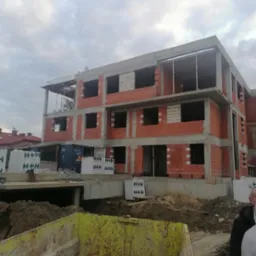 Wznoszenie trzypiętrowego budynku z czerwonej cegły H+H. Widoczne otwory okienne, szary betonowy strop i żółta skrzynia na gruz. Budowa w toku, pochmurne niebo.