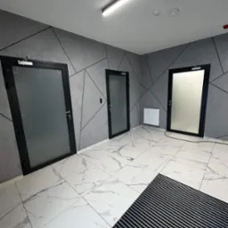 Wnętrze korytarza z trzema drzwiami z czarnymi ramami i matowym szkłem, podłoga z białych płytek imitujących marmur, ściany szare z geometrycznym wzorem.