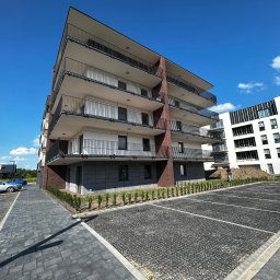 IZBUD - Nowoczesny, wielopiętrowy apartamentowiec z balkonami, wykończony cegłą i szarym kamieniem, na tle błękitnego nieba i częściowo wykończonego parkingu.