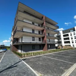 Nowoczesny, wielopiętrowy apartamentowiec z balkonami, wykończony cegłą i szarym kamieniem, na tle błękitnego nieba i częściowo wykończonego parkingu.