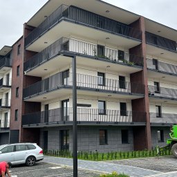 IZBUD - Nowoczesny budynek wielorodzinny z balkonami i elewacją z elementami cegły, widoczny z zewnątrz, z zaparkowanym samochodem i sprzętem budowlanym.